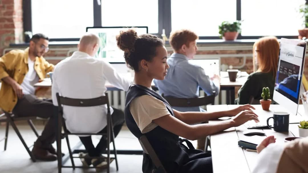 A diverse group of people working on computers in a modern office with large windows and potted plants.