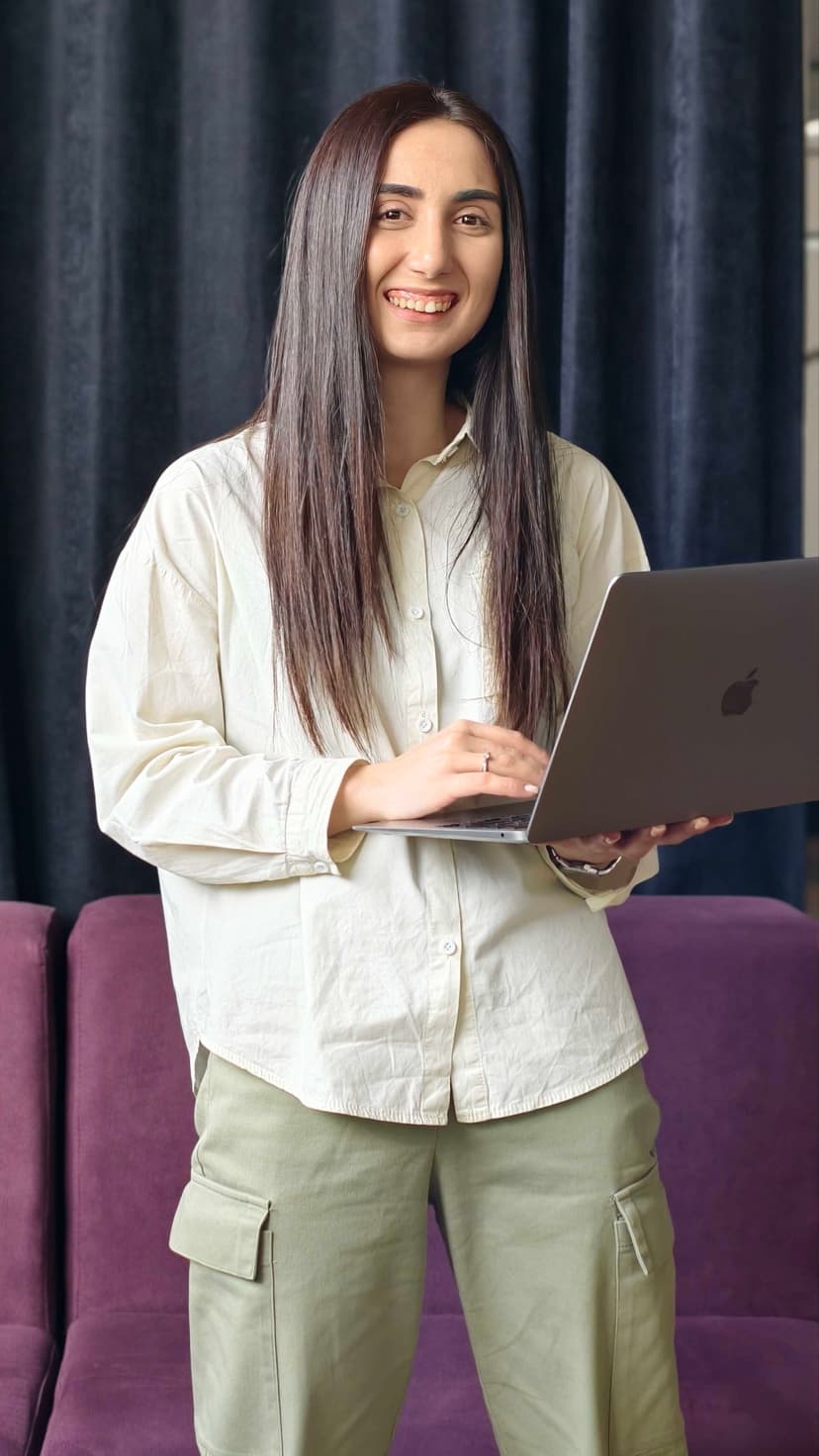 Smiling person with long hair, wearing a light shirt and green pants, holding a laptop, standing in front of dark curtains and a purple sofa.