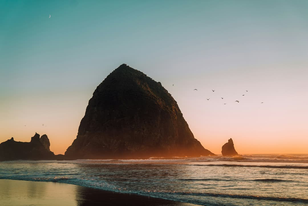 A large rock formation at the ocean's edge during sunset, with birds flying in the sky and gentle waves on the shore.